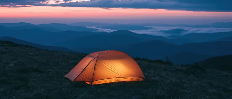 Illuminated tent on grassy mountain ridge at twilight overlooking layered valleys and fog under pink and blue sky