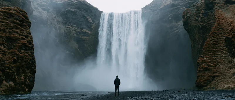 Man standing before a large waterfall in a rocky canyon, surrounded by mist and spray