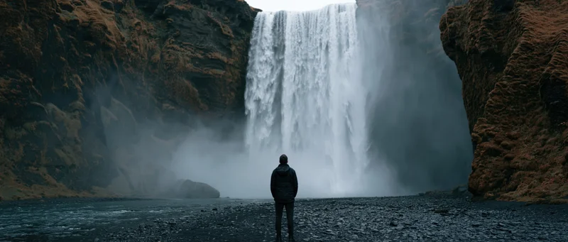 A person standing on black gravel facing a large waterfall in a rugged volcanic landscape