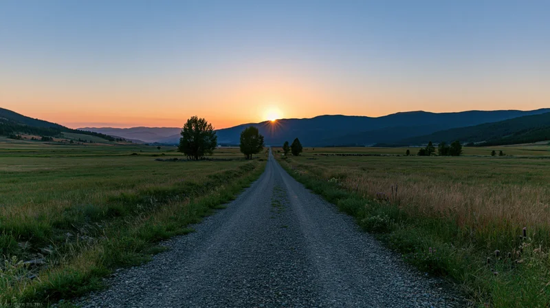 Gravel road stretching through a grassy field toward distant mountains at sunset