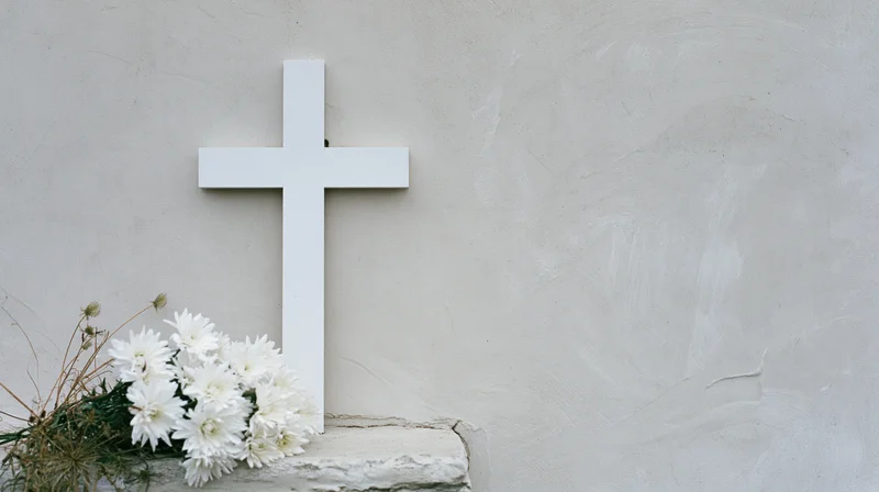 White cross on a stone grave with white chrysanthemums at its base against a plaster wall