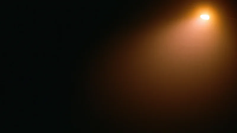 A single warm light source casting a glowing beam into dark space with soft haze and copyspace on the left