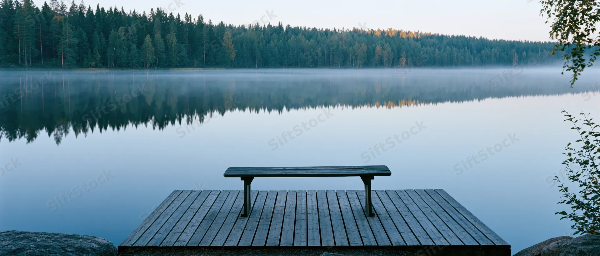 Wooden bench on a dock facing a misty lake surrounded by forest at dawn