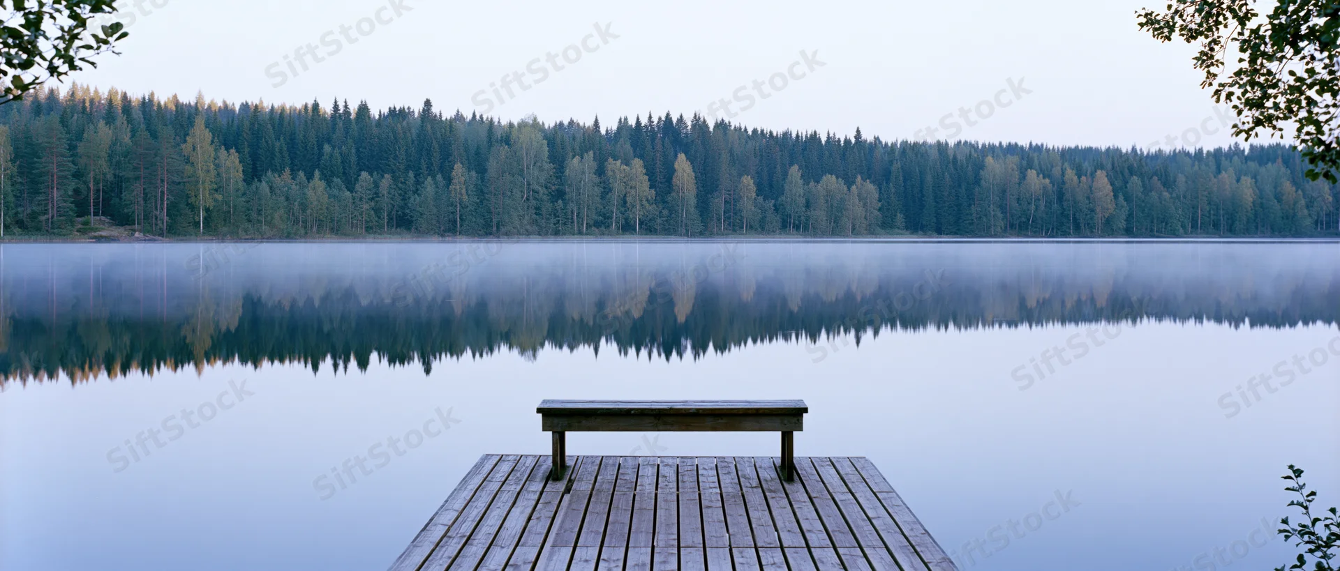 Wooden bench on a dock extending over a misty lake with forested shoreline reflected in calm water