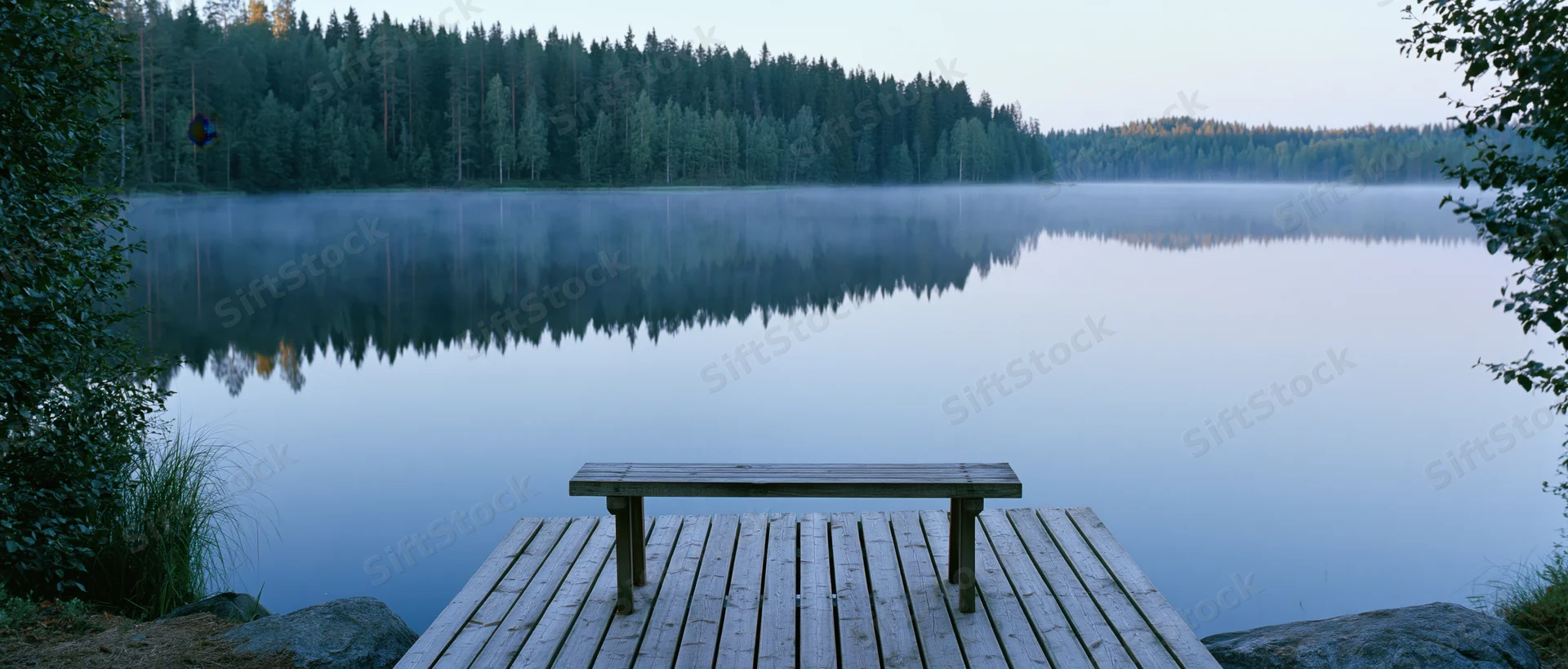 Wooden bench on a dock overlooking a misty lake surrounded by dense forest at dawn