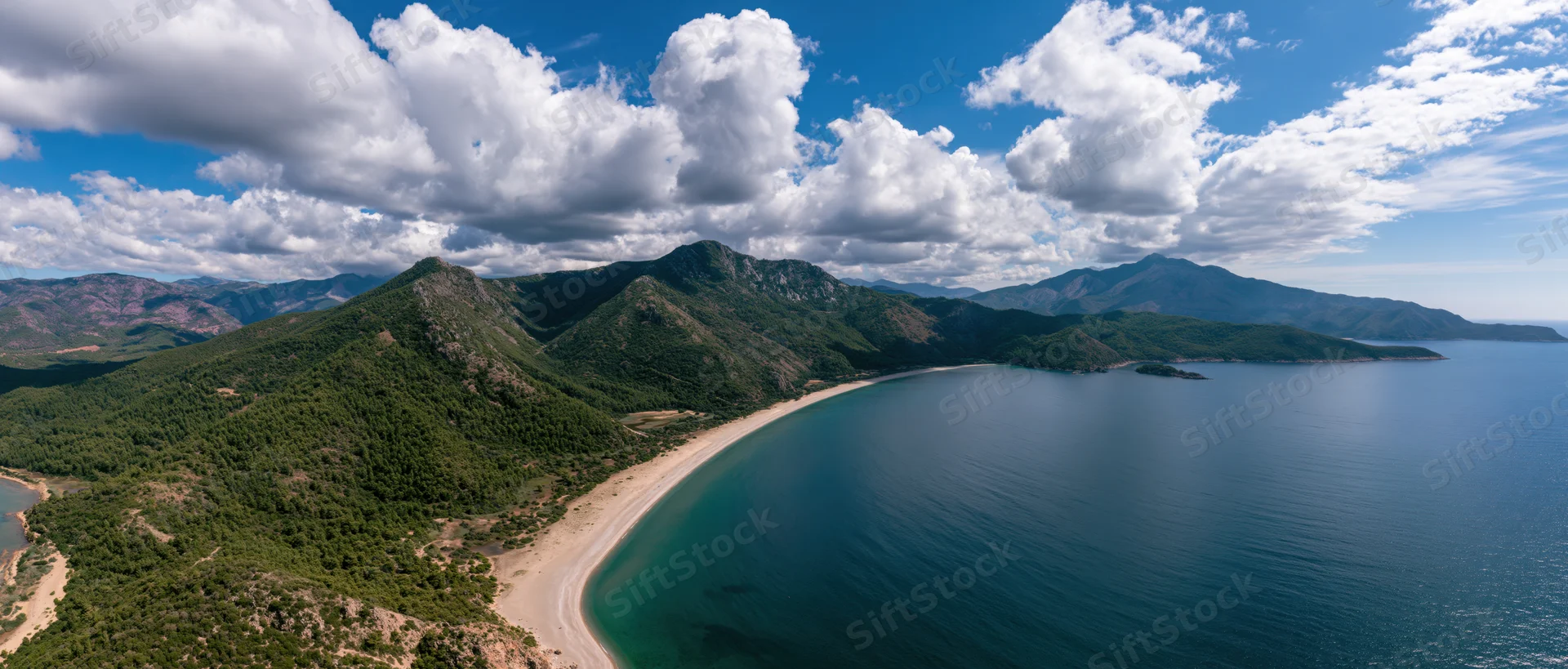 Aerial view of a sandy beach curving along a turquoise sea bordered by green mountains under a blue sky with white clouds