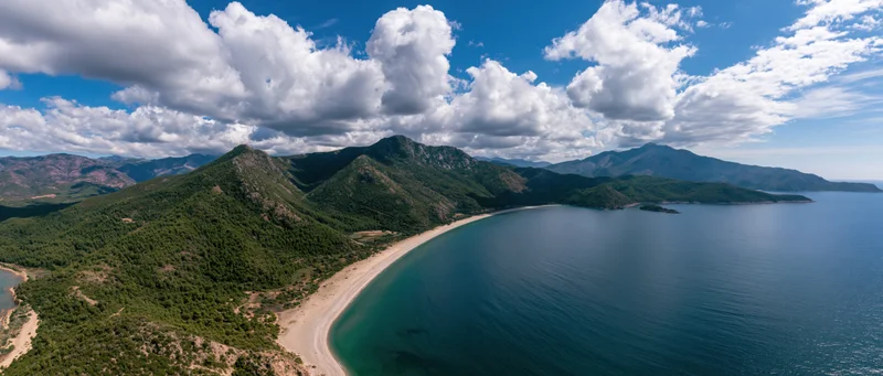 Aerial view of a sandy beach curving along a turquoise sea bordered by green mountains under a blue sky with white clouds
