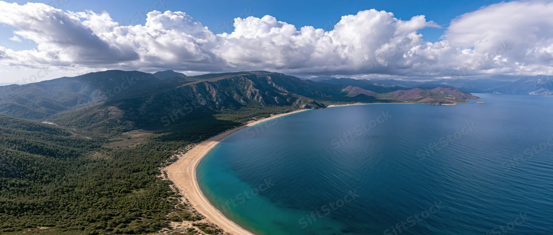 Aerial view of a curved sandy beach bordered by green mountains and deep blue sea under a cloudy sky