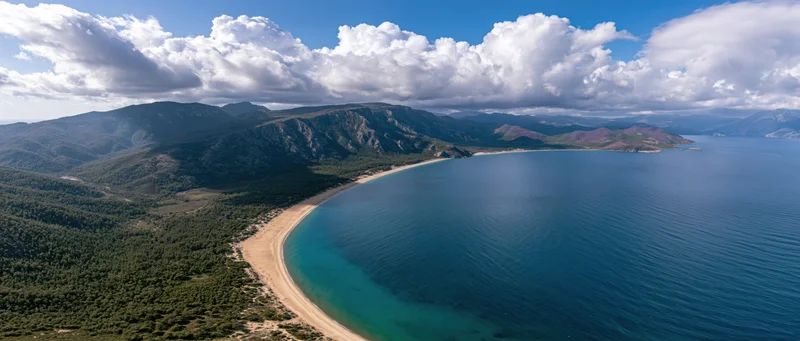 Aerial view of a curved sandy beach bordered by green mountains and deep blue sea under a cloudy sky