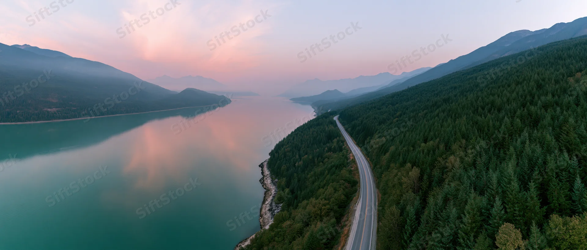 Aerial view of a winding road along a turquoise lake surrounded by forested mountains at sunset