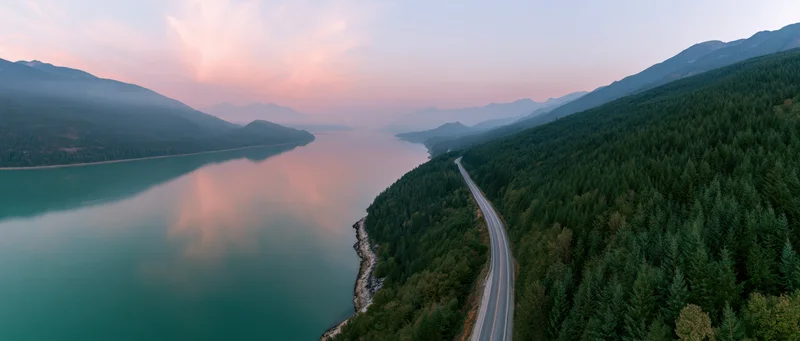 Aerial view of a winding road along a turquoise lake surrounded by forested mountains at sunset