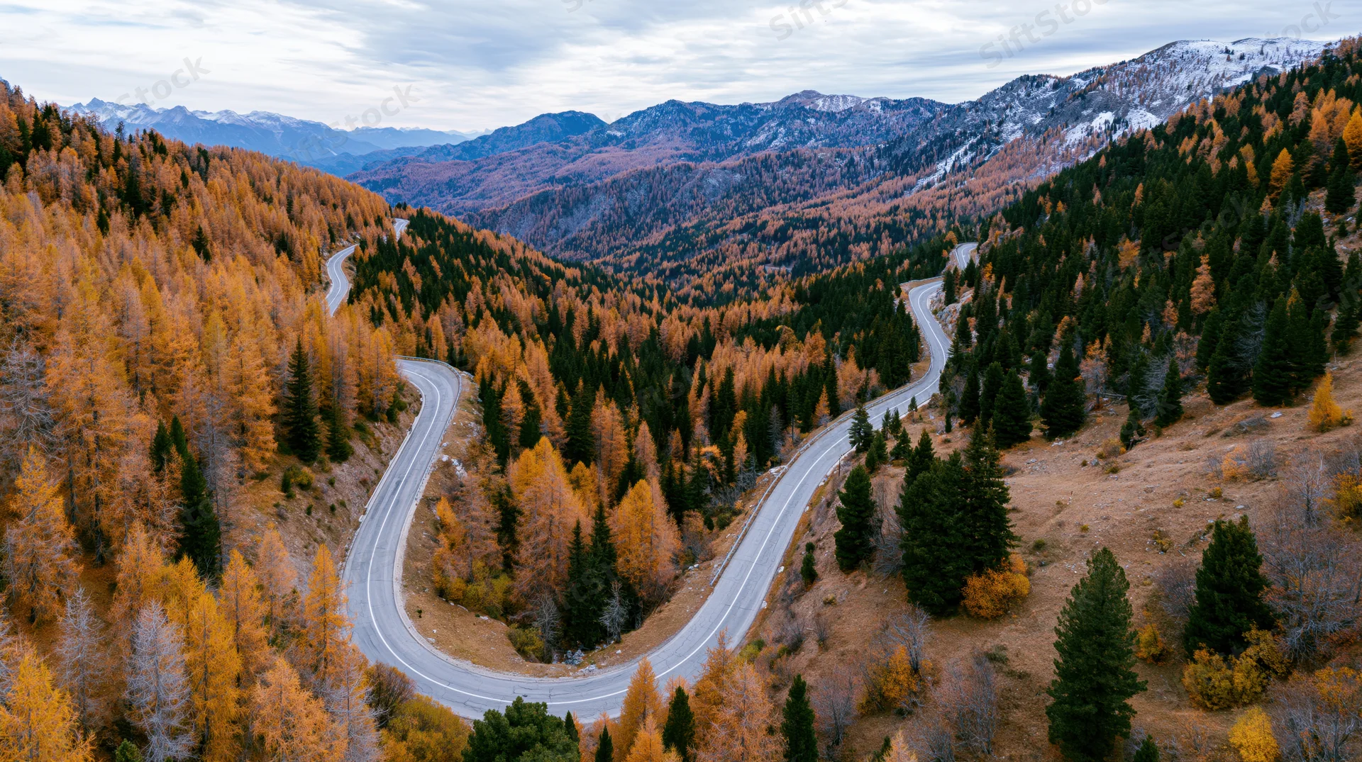 Winding asphalt road curves through autumn forested mountain valley with snow-capped peaks in distance