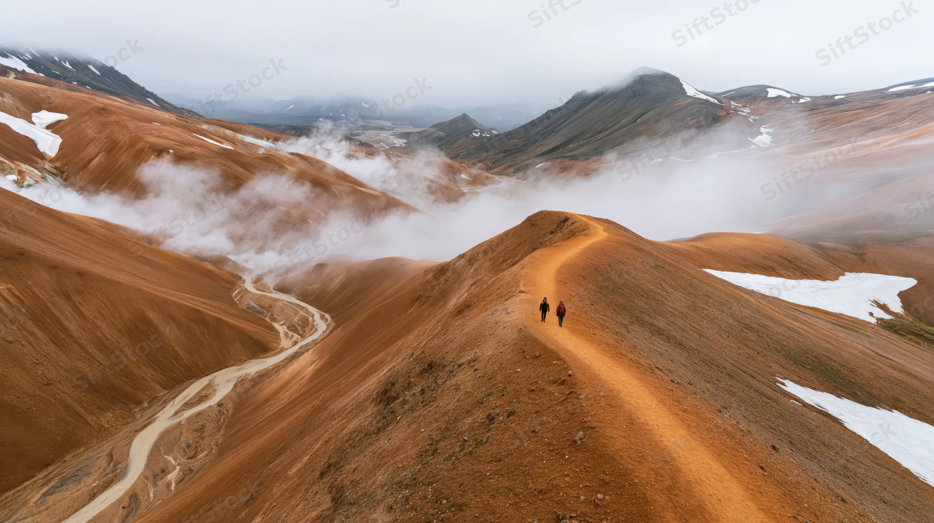 Two hikers walking along a narrow mountain ridge through foggy alpine terrain with snow patches and winding trails below
