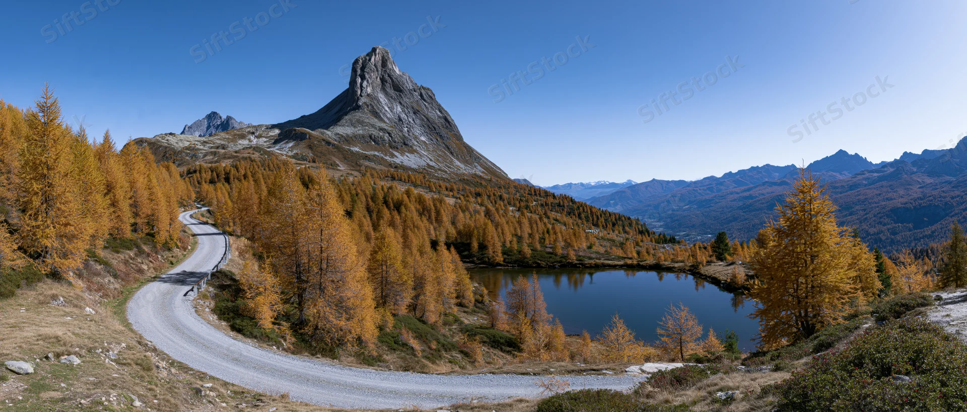 Winding gravel road through autumn larch forest beside alpine lake with mountain peak in background