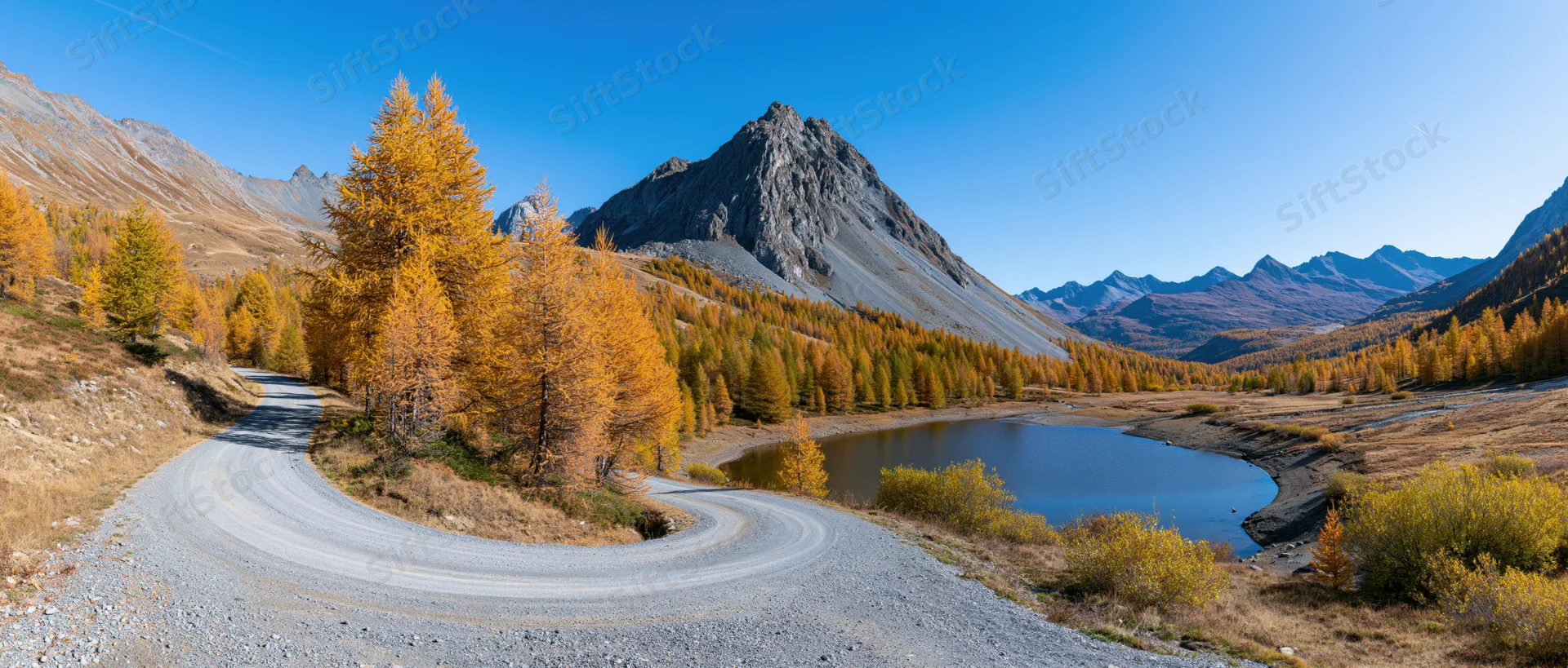 Winding gravel road curves through autumn larch forest beside alpine lake in mountain valley