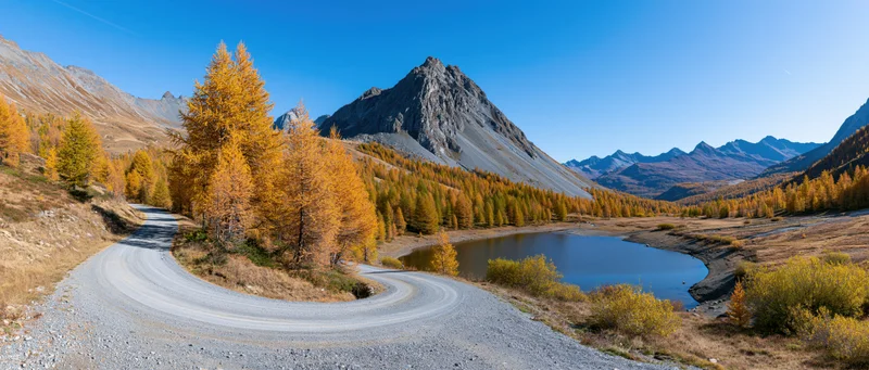 Winding gravel road curves through autumn larch forest beside alpine lake in mountain valley