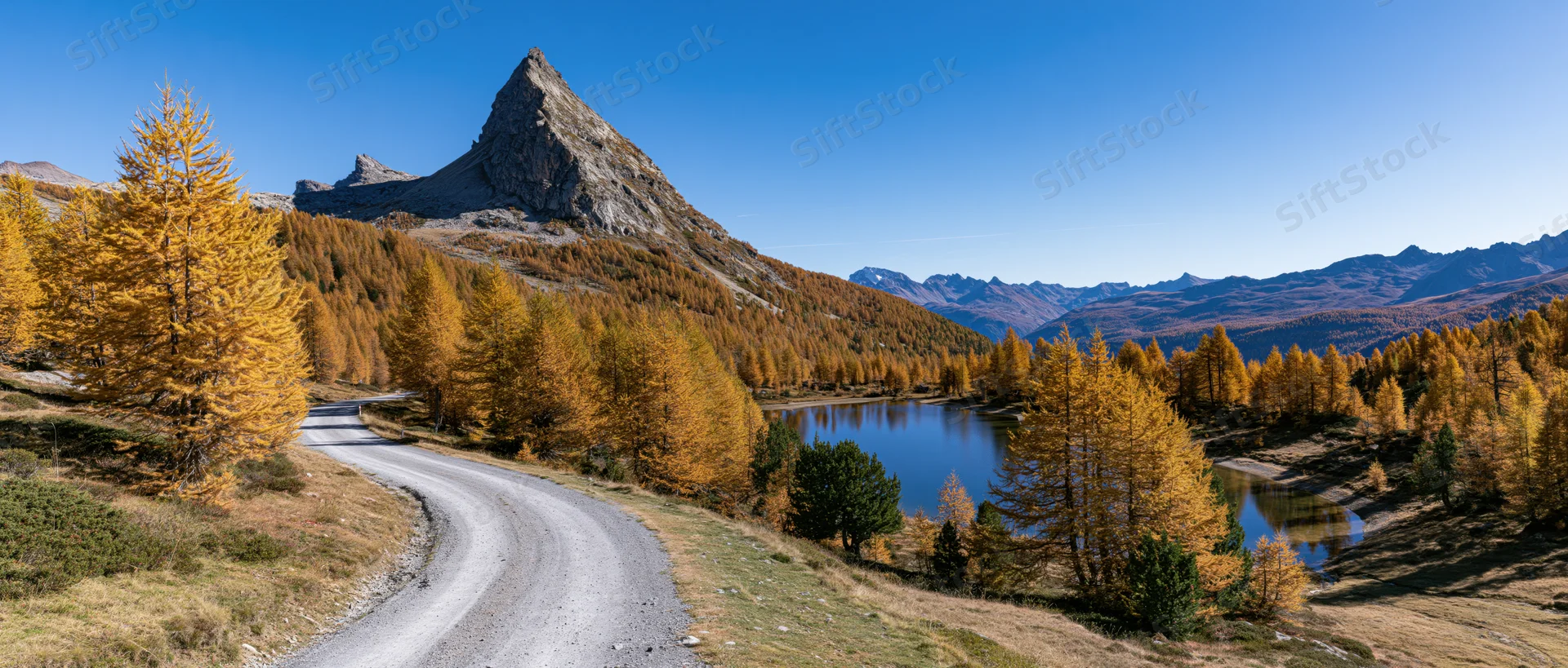 Winding gravel road through golden larch forest beside alpine lake under sharp mountain peak