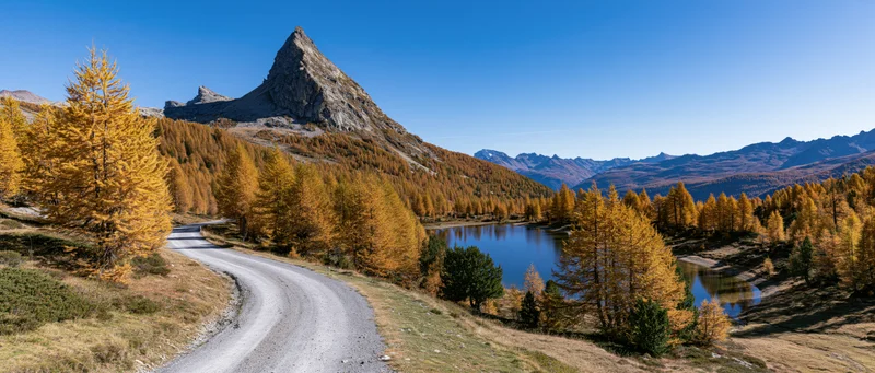 Winding gravel road through golden larch forest beside alpine lake under sharp mountain peak