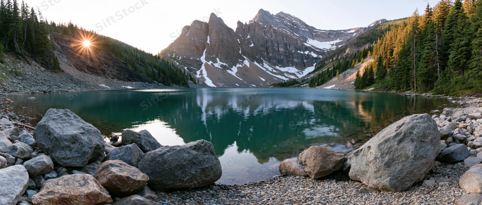 Mountain lake reflecting peaks at sunrise surrounded by pine forest and boulders