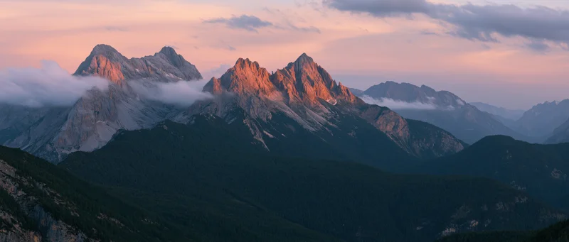 Rugged mountain peaks at dusk with clouds drifting through valleys and forested slopes below
