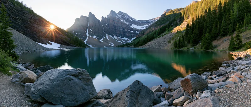 Alpine lake reflecting snow-capped mountains at sunrise surrounded by evergreen forest and rocky shore