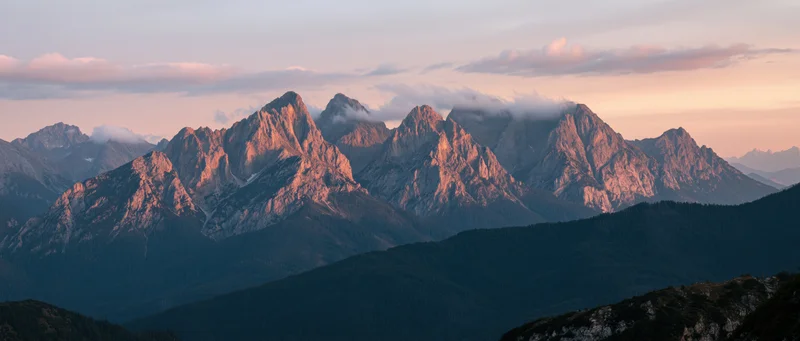 Rugged mountain peaks bathed in alpenglow light at sunset with forested slopes below