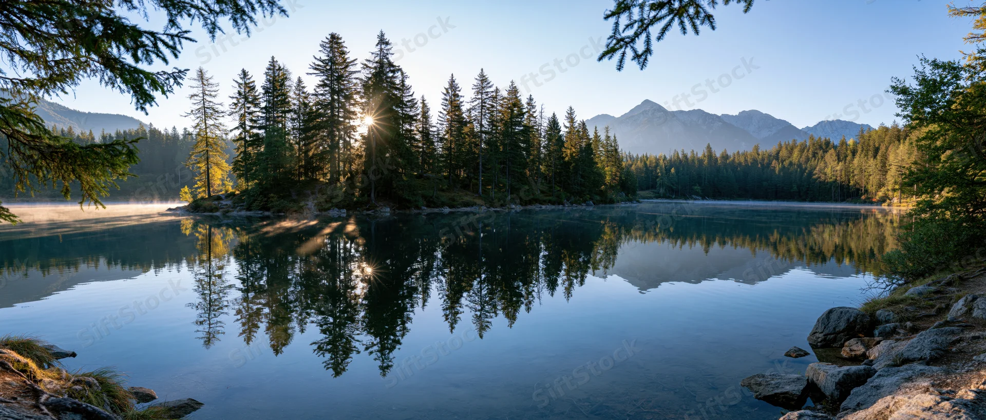 Calm lake reflecting pine trees and mountains at sunrise with mist rising from the water