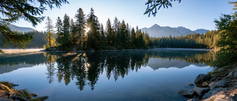 Calm lake reflecting pine trees and mountains at sunrise with mist rising from the water