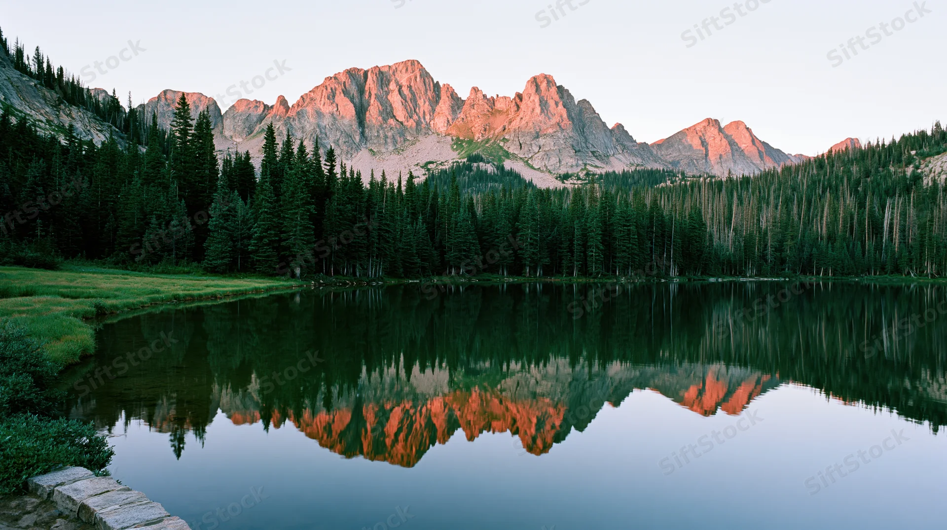 Rugged mountain peaks reflected in calm lake surrounded by dense evergreen forest at sunrise
