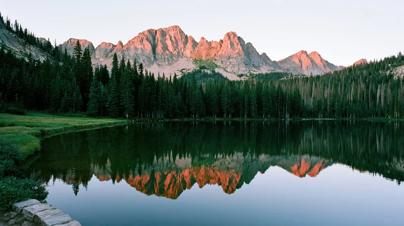 Rugged mountain peaks reflected in calm lake surrounded by dense evergreen forest at sunrise