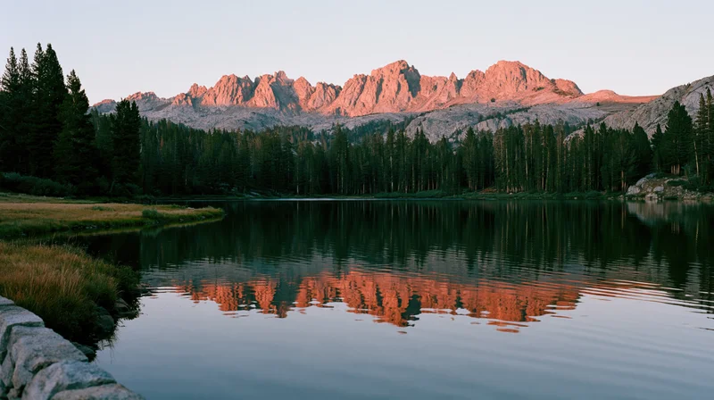 Mountain range reflected in calm lake surrounded by pine forest at sunset