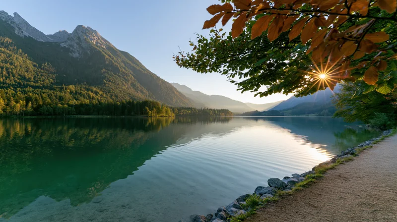 Calm lake reflecting mountains and forest at sunrise along a gravel path with autumn leaves overhead