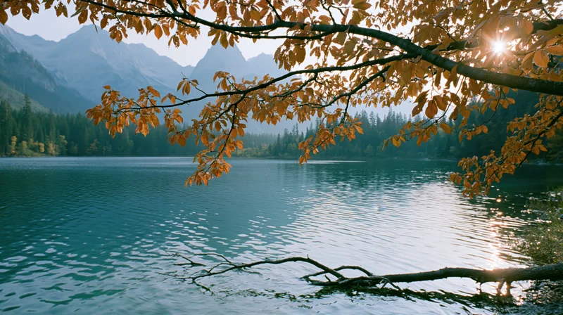 Autumn tree branches with golden leaves over a calm lake reflecting sunlight and distant mountains