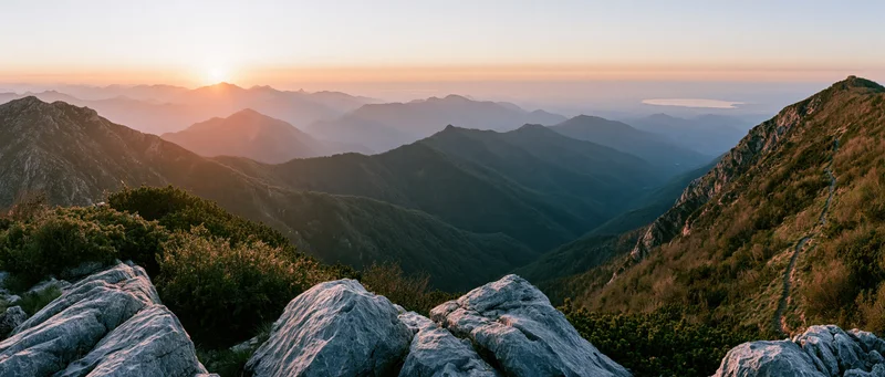 Sunrise over layered mountain ridges with rocky foreground and forested valleys