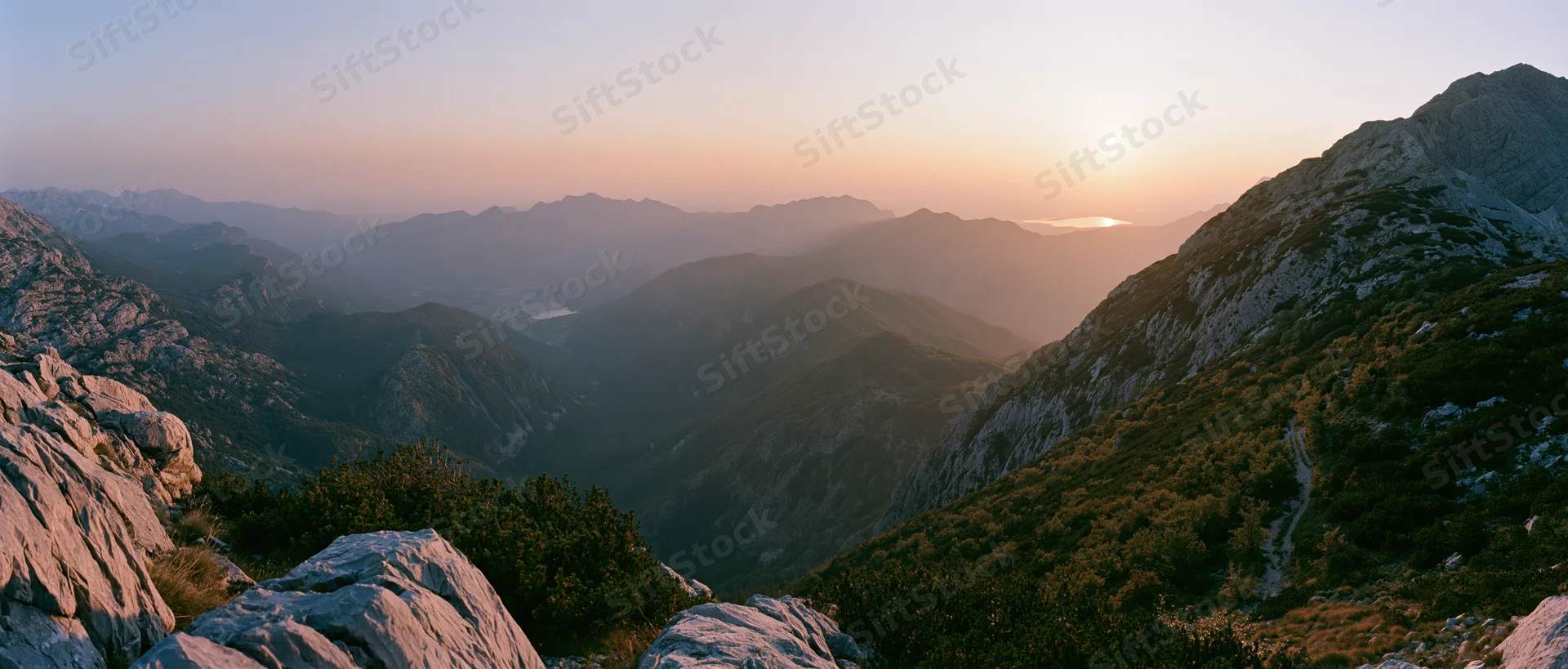 Mountain range at sunset viewed from a high ridge overlooking a valley with forested slopes and rocky outcrops