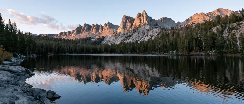 Mountain peaks reflected in calm lake surrounded by pine forest at sunset
