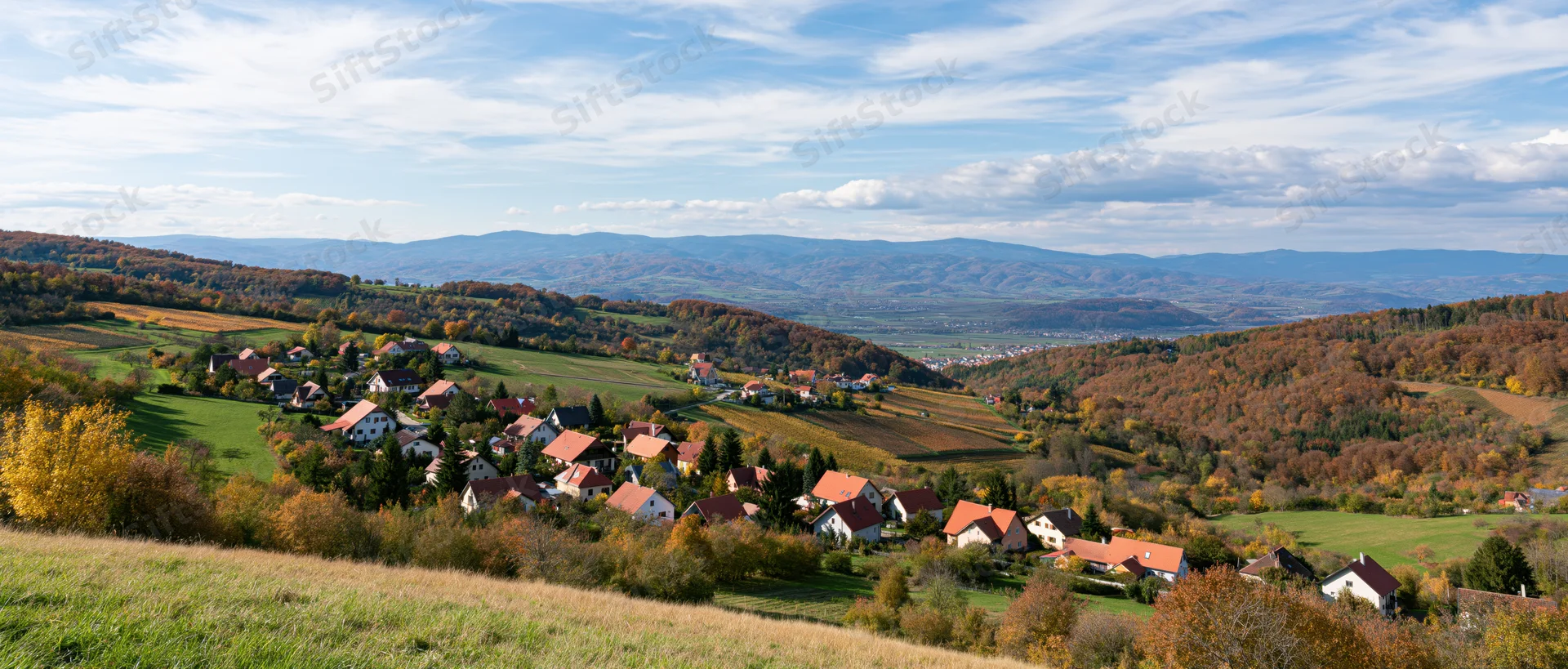 Village nestled among autumn-colored hills and fields under a blue sky with scattered clouds