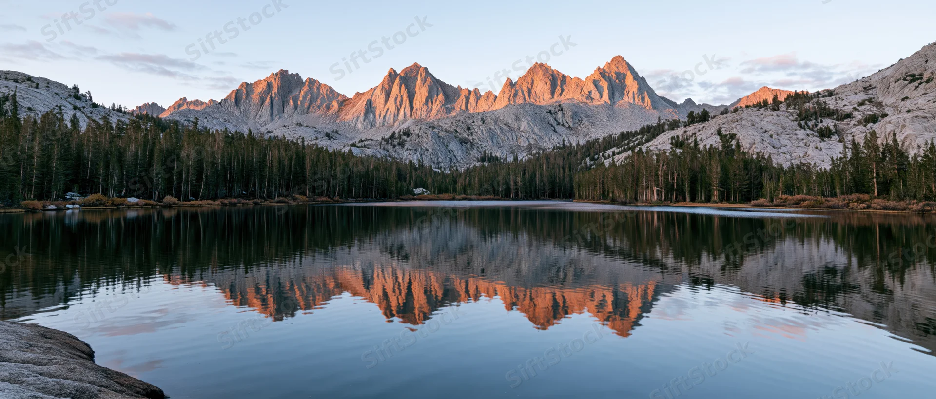 Mountain range reflected in calm lake surrounded by evergreen forest at sunset