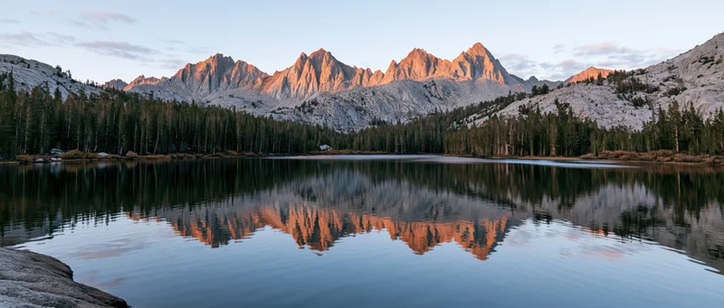 Mountain range reflected in calm lake surrounded by evergreen forest at sunset