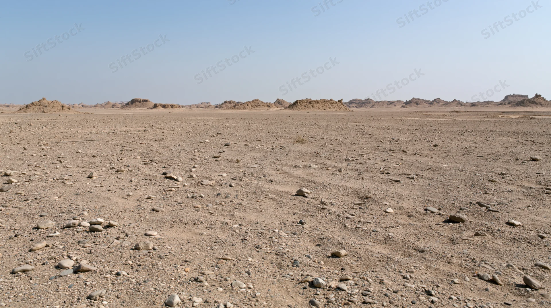 Arid rocky desert landscape with scattered stones and distant mounds under clear blue sky