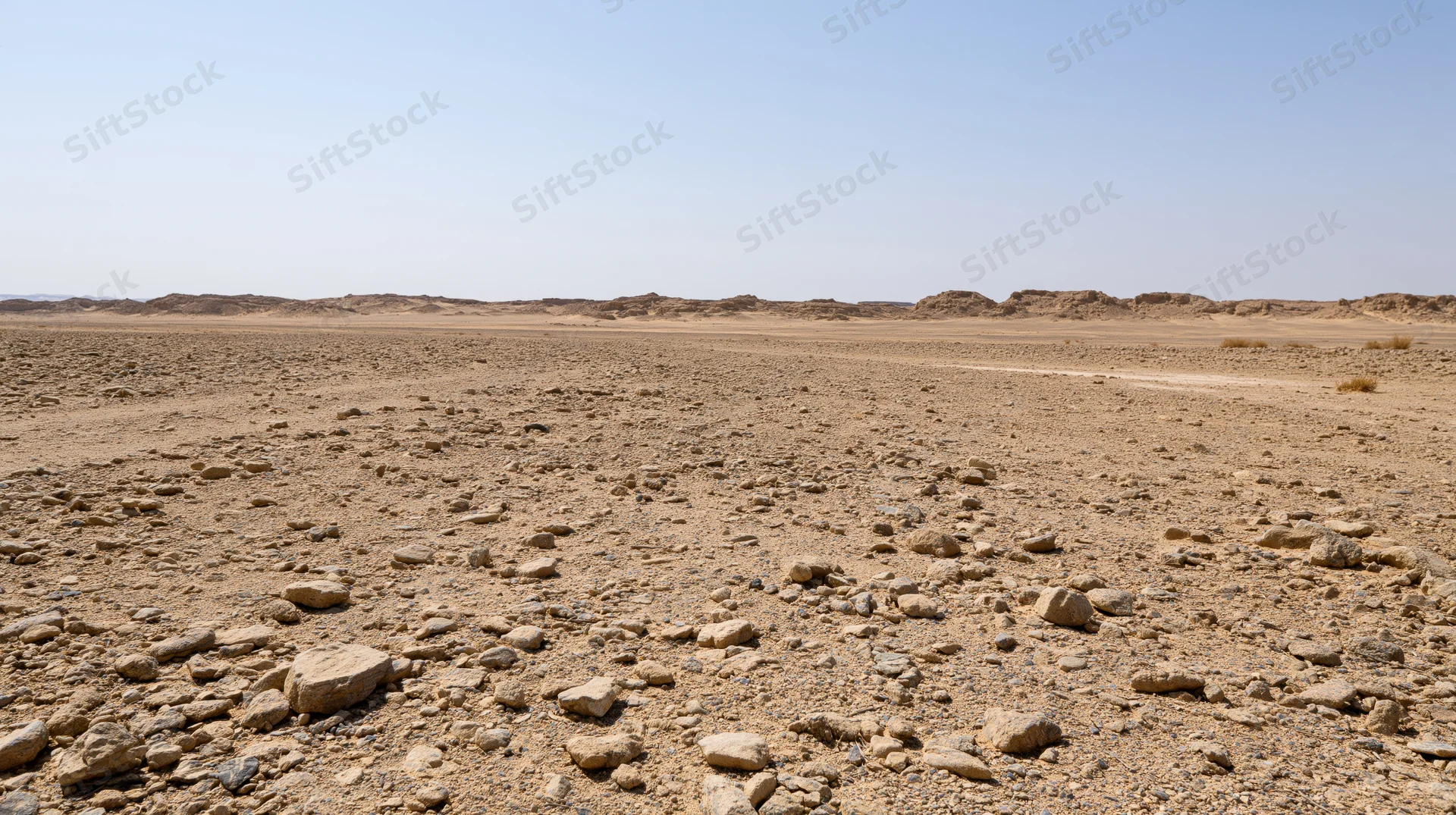 Rocky desert terrain under clear blue sky with distant plateaus and scattered stones