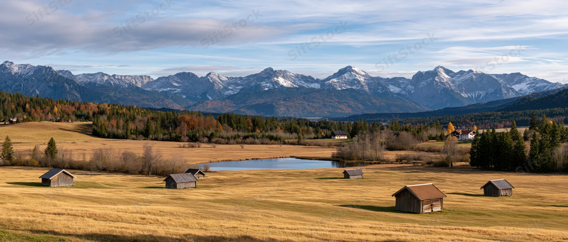Wooden barns scattered across a golden field with snow-capped mountains in the background during autumn
