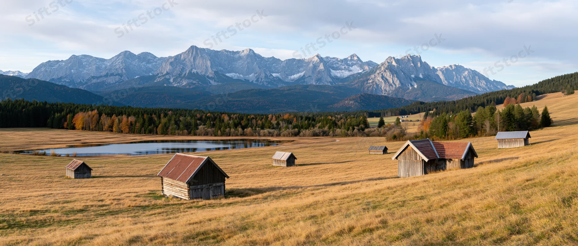 Wooden barns scattered across a golden meadow with alpine mountains in the background during autumn
