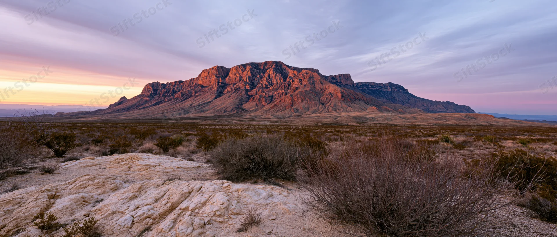 Rocky mountain formation at sunset in a desert landscape with sparse shrubbery and layered rock formations under a pastel sky