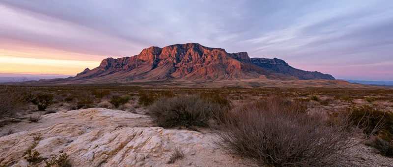 Rocky mountain formation at sunset in a desert landscape with sparse shrubbery and layered rock formations under a pastel sky