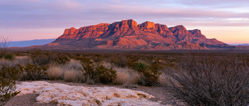 Red rock mountain formation at sunset in desert landscape with sparse vegetation and distant horizon