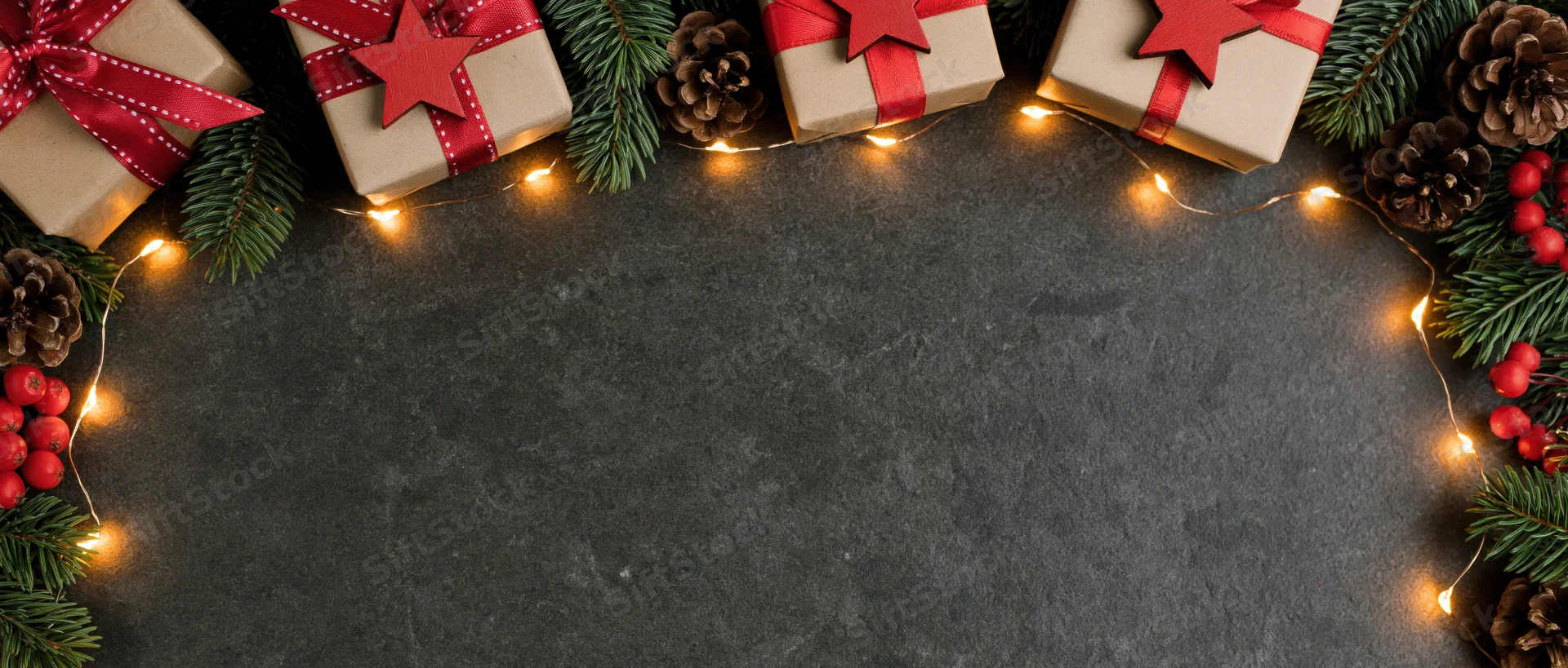 Christmas gifts wrapped in kraft paper with red ribbons and stars arranged with pine branches, pinecones, berries and string lights along the top edge of a dark stone surface, leaving copyspace below