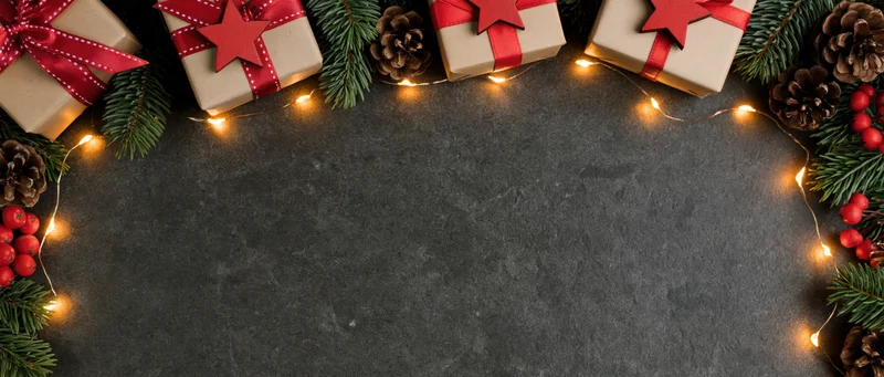 Christmas gifts wrapped in kraft paper with red ribbons and stars arranged with pine branches, pinecones, berries and string lights along the top edge of a dark stone surface, leaving copyspace below