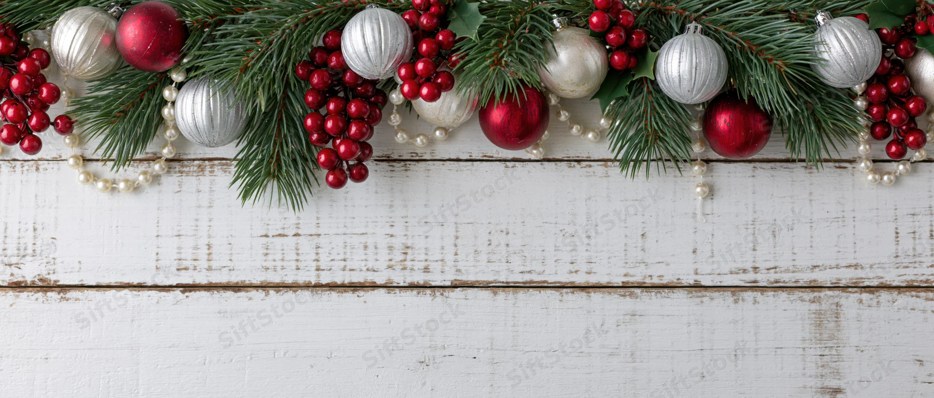 Christmas garland with pine branches, red and silver ornaments, and berries arranged along white wooden surface, leaving copyspace below