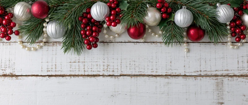 Christmas garland with pine branches, red and silver ornaments, and berries arranged along white wooden surface, leaving copyspace below